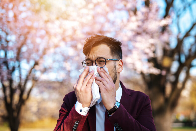Ein Mann in einem Anzug benutzt ein Taschentuch, um sich die Nase zu putzen, vor blühenden Bäumen im Hintergrund.