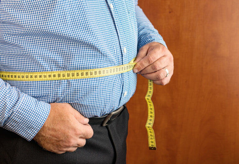 adipositas.jpg Mann misst seinen Bauchumfang mit einem gelben Maßband, trägt ein kariertes Hemd und dunkle Hose.