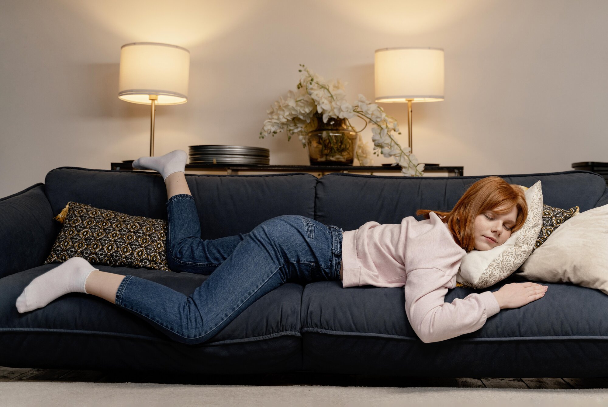powernap.jpg Eine junge Frau mit roten Haaren liegt entspannt auf einem blauen Sofa, umgeben von Kissen und Lampen.