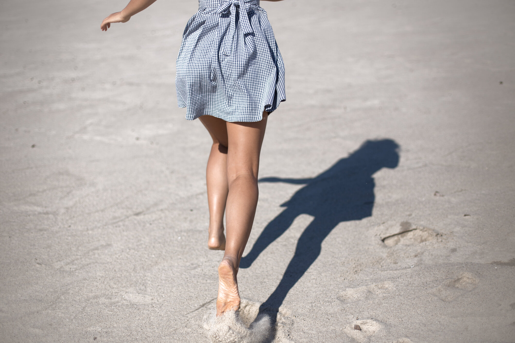 Beine.jpg Eine Person in einem karierten Kleid läuft barfuß über einen sandigen Strand, während ihr Schatten sichtbar ist.