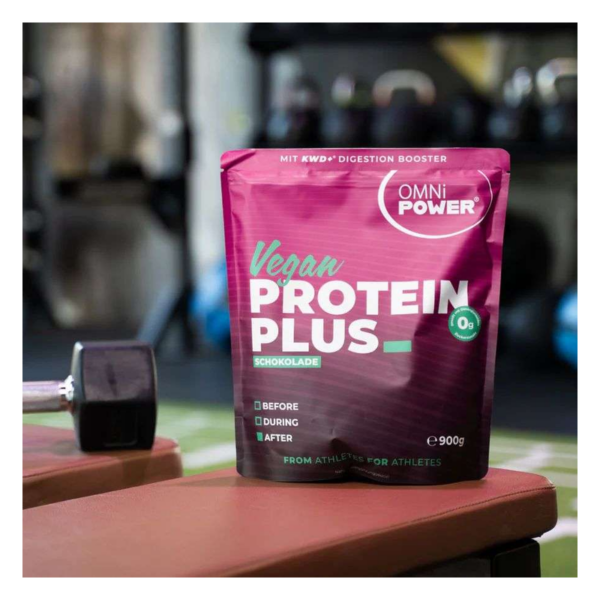 Eine violette Tüte mit veganem Proteinpulver in Schokoladengeschmack, platziert auf einer Trainingsbank in einem Fitnessstudio.