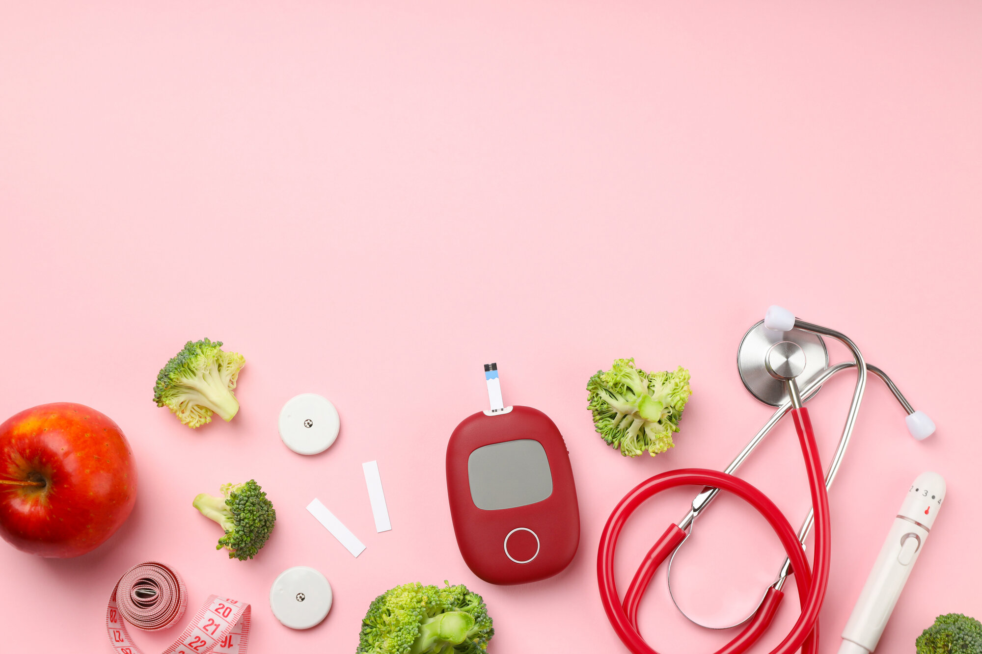 Blutzucker spitzen.jpeg Gesunde Lebensmittel und medizinische Geräte auf rosa Hintergrund, darunter Apfel, Brokkoli, Blutzuckermessgerät und Stethoskop.
