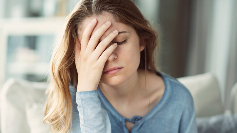 Junge Frau mit langen Haaren und blauem Oberteil, die frustriert ihre Hand an die Stirn legt, im Innenraum sitzend.