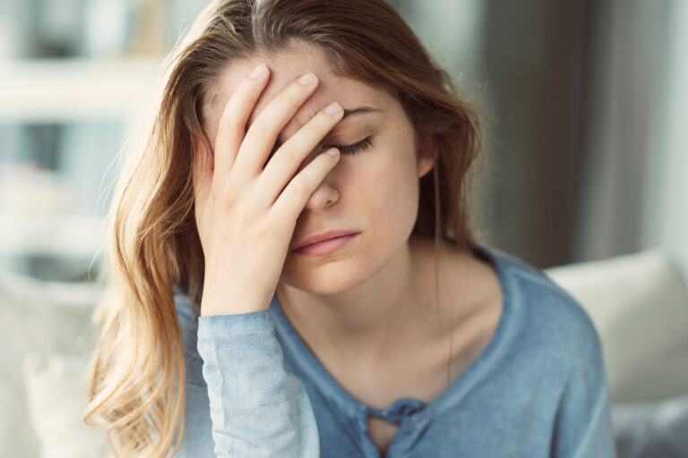 Junge Frau mit langen Haaren und blauem Oberteil, die frustriert ihre Hand an die Stirn legt, im Innenraum sitzend.